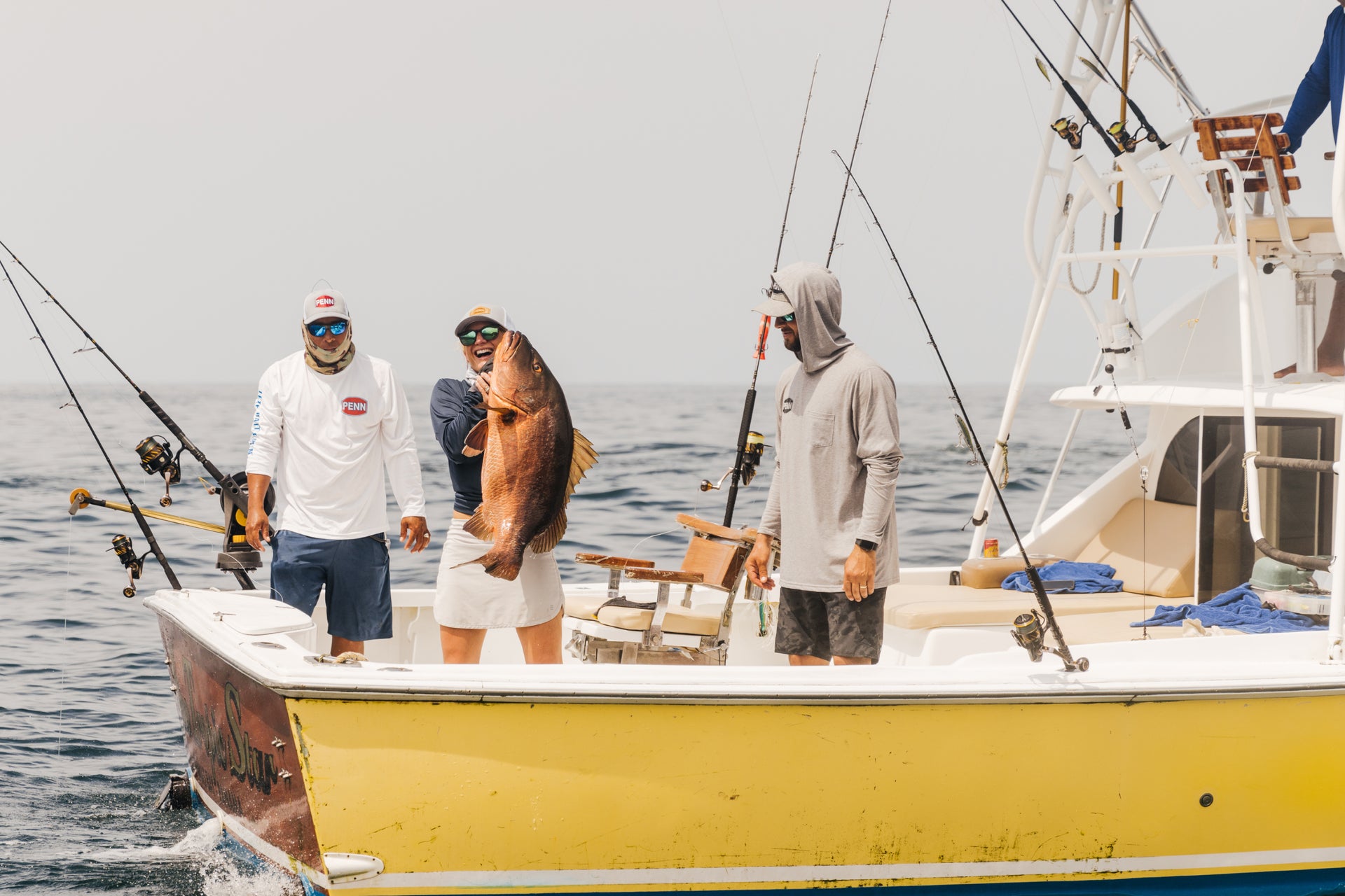Three men on a boat with one holding a large fish, wearing hats and casual clothing.