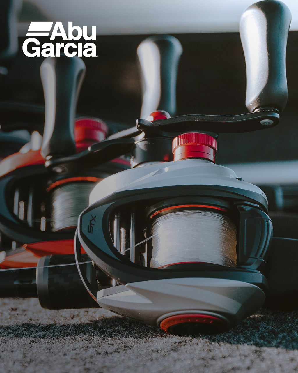 A close-up of an Abu Garcia baitcasting fishing reel with a black and silver body, red accents, and white line spool, resting on sand. The Abu Garcia logo is visible in the top left corner.